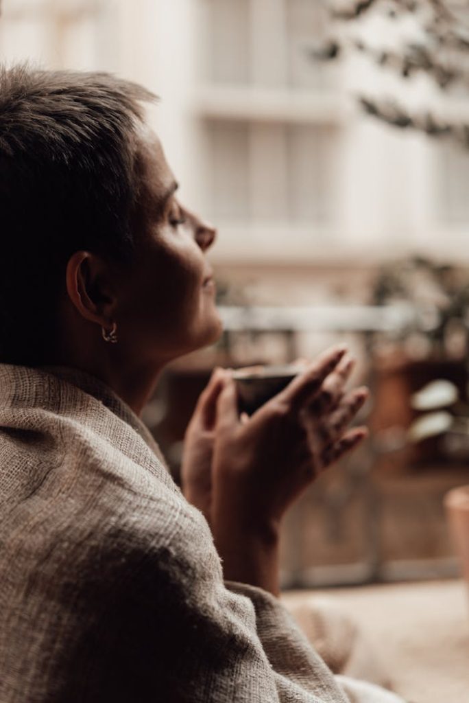 crop-dreamy-woman-enjoying-coffee-aroma-on-balcony-6842245 A peaceful moment of a woman savoring a hot drink, wrapped in a blanket with eyes closed, exuding tranquility.